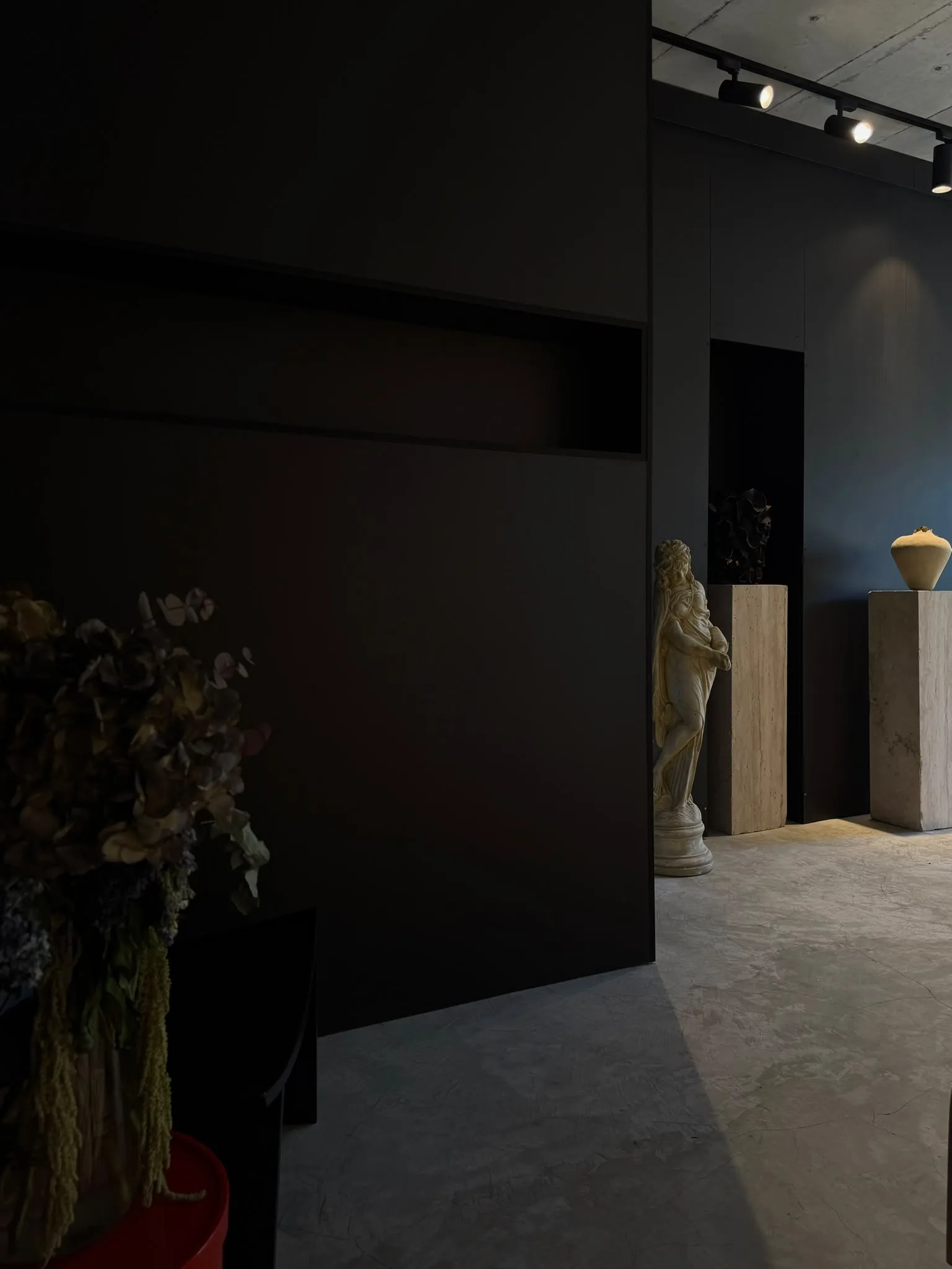 Dried flowers in a dim corner; beyond, a plaster figure and a vessel on a plinth