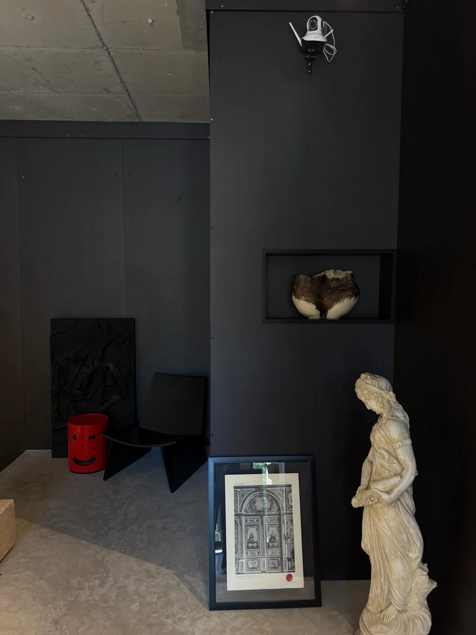 A ceramic vessel in a niche in the black wall, a plaster figure and a framed print in front