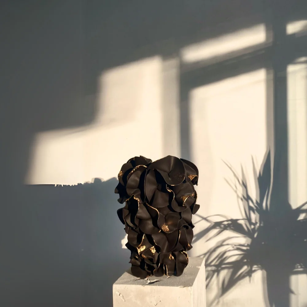 Petal: a ruffled black vessel flecked with gold on a white plinth, window light and a plant's shadow on the wall behind it